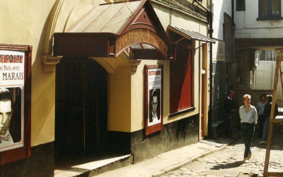 Exterior shot of City Varieties box office alongside a cobbled street, the sign reading 'Theatre Royal'. Posters on the building depict a man's face with the name 'Alain Marais'.