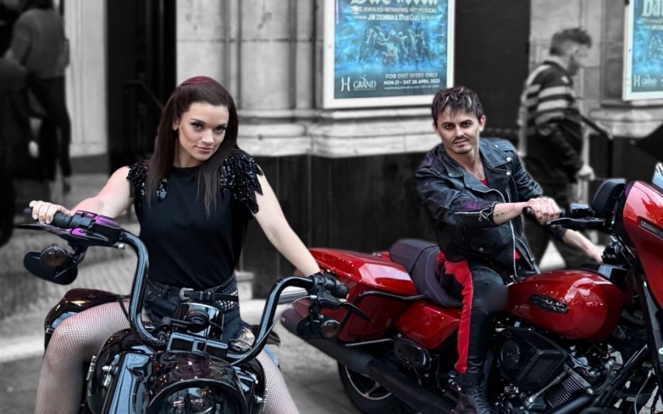 Two cast members from Bat Out of Hell pose on motorcycles outside Leeds Grand Theatre
