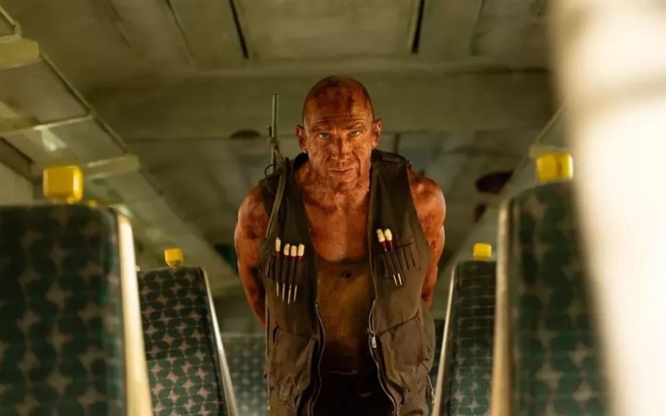 Dirty and bloodied Ralph Fiennes wearing a camo jacket, containing darts and a rifle on his back, standing on an empty train.