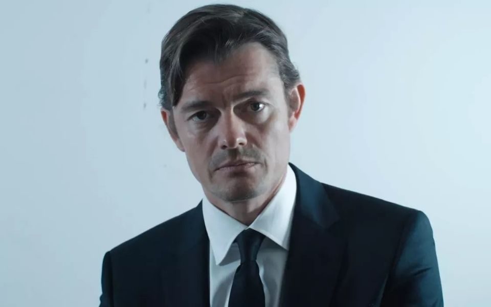 A man in a suit looks deadpan at the camera, with muted tones.