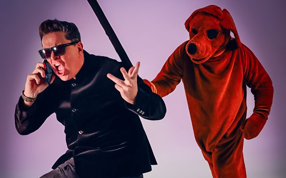 Dom Joly comically shouts into a mobile phone, wearing a shiny black jacket and sunglasses. A person in a reddish dog suit is behind him with a baton