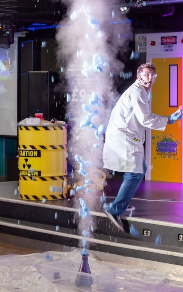 From a beaker on the ground, blue foam erupts upwards accompanied by smoke, as a cast member in a lab-coat retreats upstage.