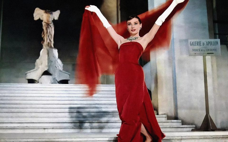 Audrey Hepburn poses with outstretched arms at the bottom of some stone steps, in a red dress, white gloves and red shawl.
