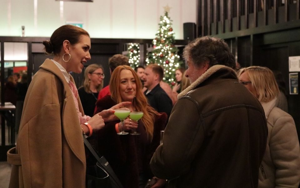 Audience members enjoying green cocktails, with a glowing Christmas tree behind them