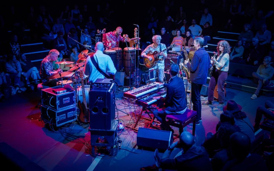 The band Hejira play to a crowd surrounding them 'in the round', lit by blue and red stage lighting.