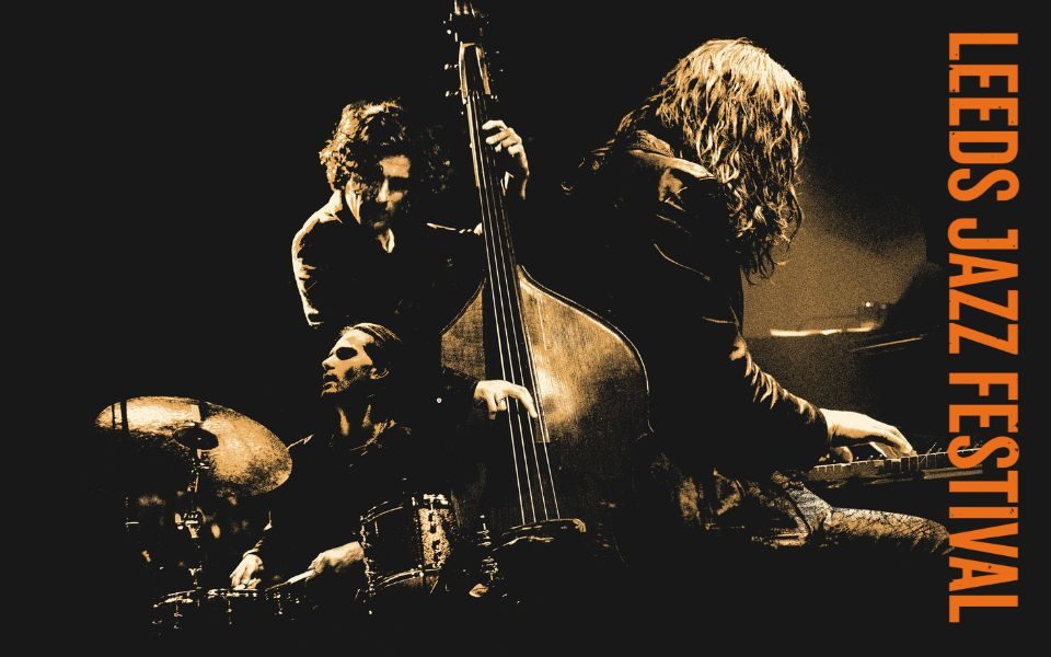 Dramatically lit in high contrast shadows, a central double bass player is flanked either side by a drummer and long-haired pianist. Text reads: Leeds Jazz Festival.