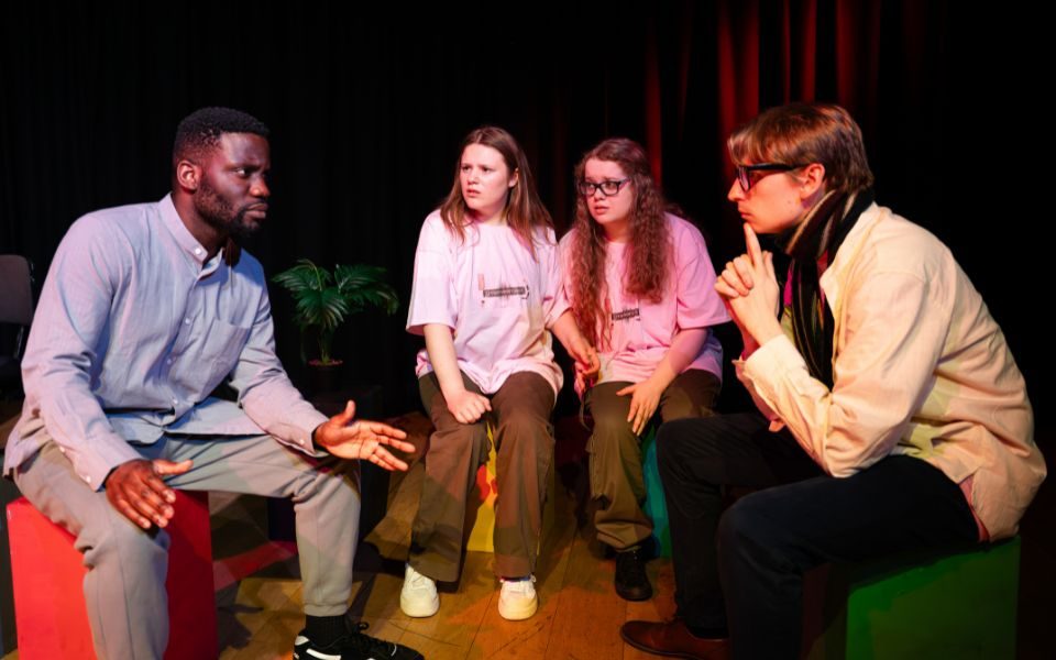 Actors sit, huddled around, focused in a deep and concerned conversation on a dimly lit stage.