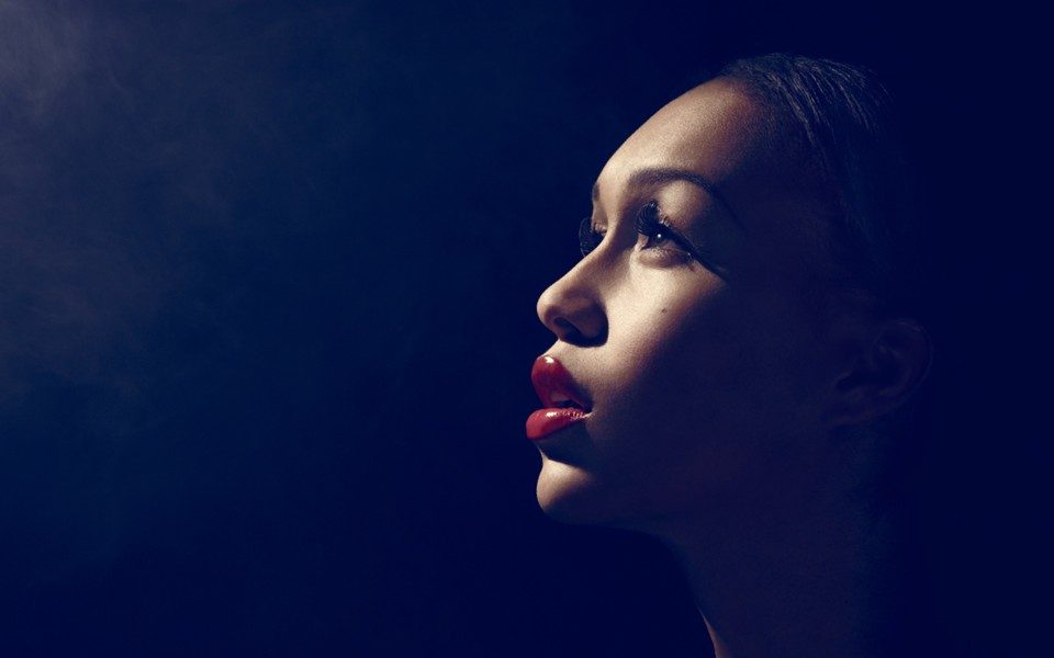 Lit by a solitary spotlight against a dark, hazy backdrop, Rebecca Ferguson looks into the distance with elegant makeup and red lipstick.