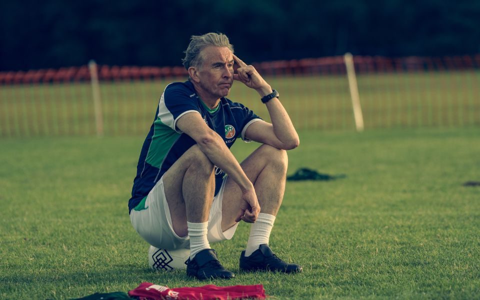 Wearing an Irish football kit, Steve Coogan squats on a football, with a hand raised to his head, at a training ground.
