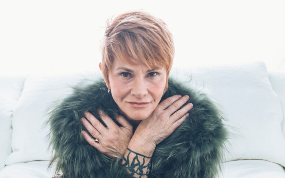 Shawn Colvin smirks with hands across her chest, wearing a furry green jacket, sat on a bright white bed.