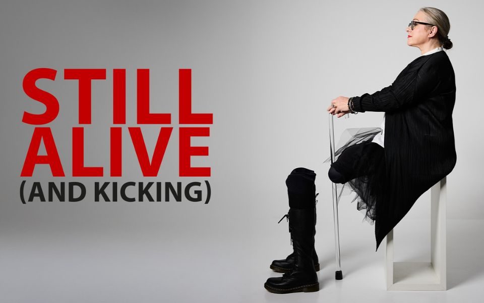 Gill Hicks sat in profile on a stool, holding a stick with her head raised. A pair of boots are positioned in front of her. Text reads: Still Alive (and Kicking).