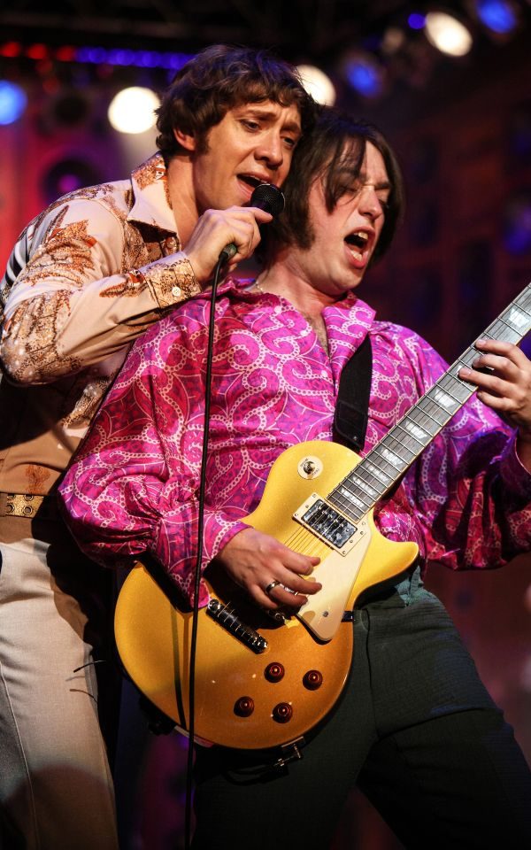 A man singing into a microphone behind a man in a pink shirt playing the guitar.