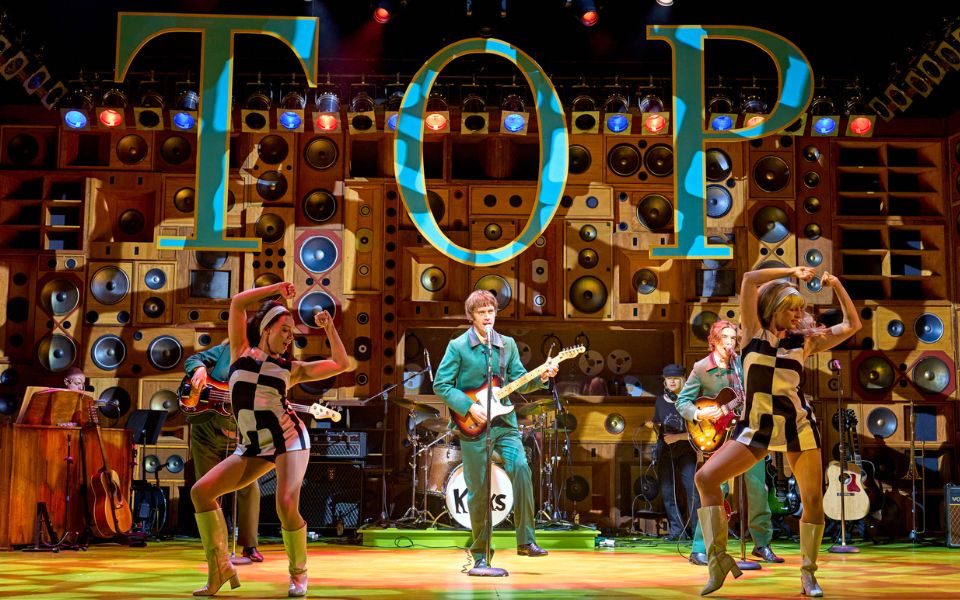 The Kinks band onstage with dancers wearing black and white 60s dresses. Above them is a big sign reading 'TOP'.
