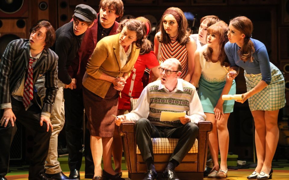 A crowd gathered around a man in an armchair holding a piece of paper. They all crane their heads in the same direction to look at something intently.