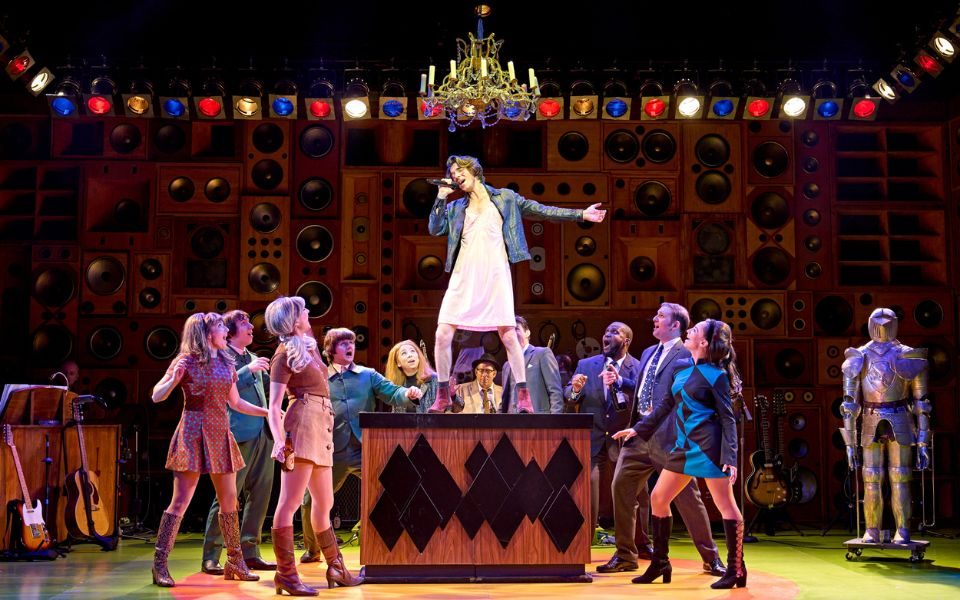 Oliver Hoare dressed in a dress and leather jacket singing into a microphone while standing on top of a cabinet as a crowd look up at him in awe.