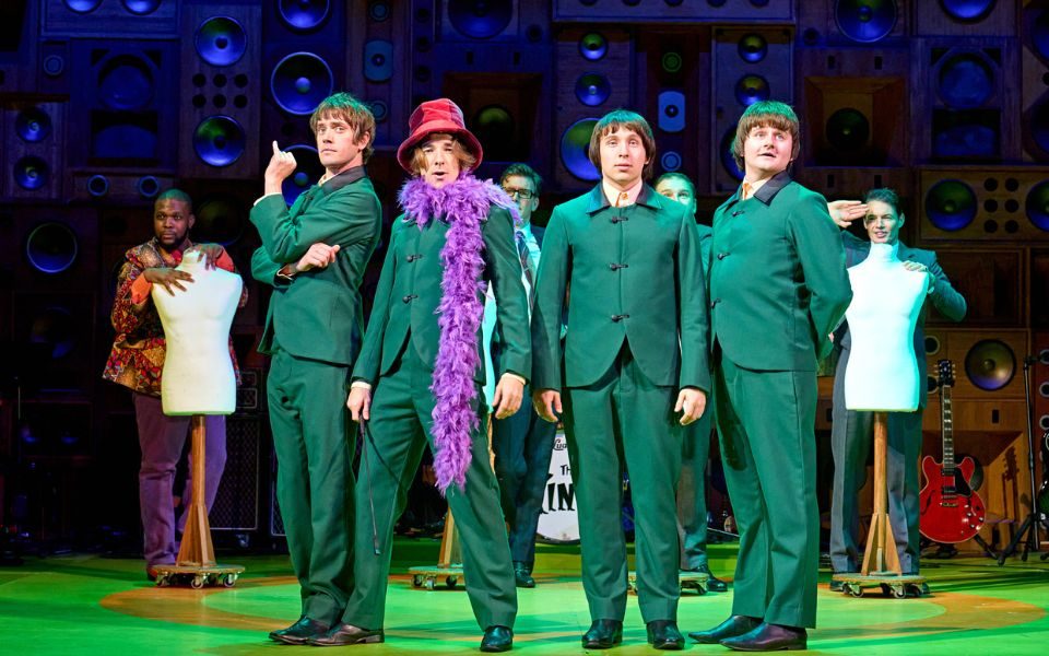 Actors playing The Kinks stand in a line wearing matching green suits. One of them wears a red hat and purple feather boa.