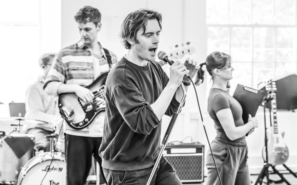 A black and white photo of Danny Horn singing into a microphone in rehearsals for Sunny Afternoon.