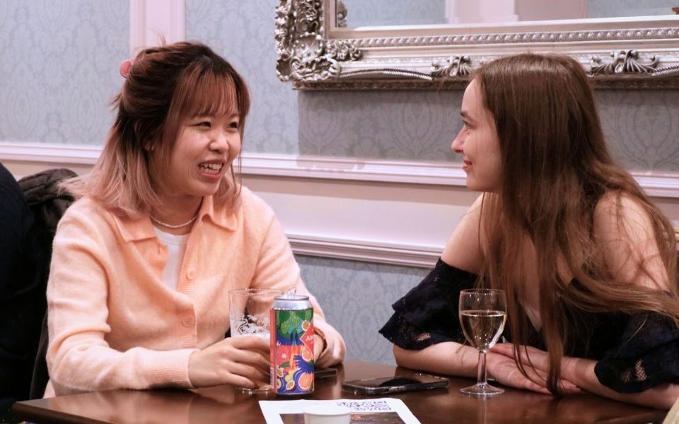 Two young people sit smiling and chatting, enjoying a glass of wine and a beer.
