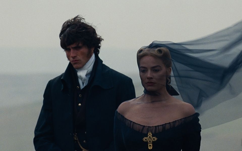 Jacob Elordi and Margot Robbie in period costume, wearing a dark veil blowing in the wind, set against a rural backdrop