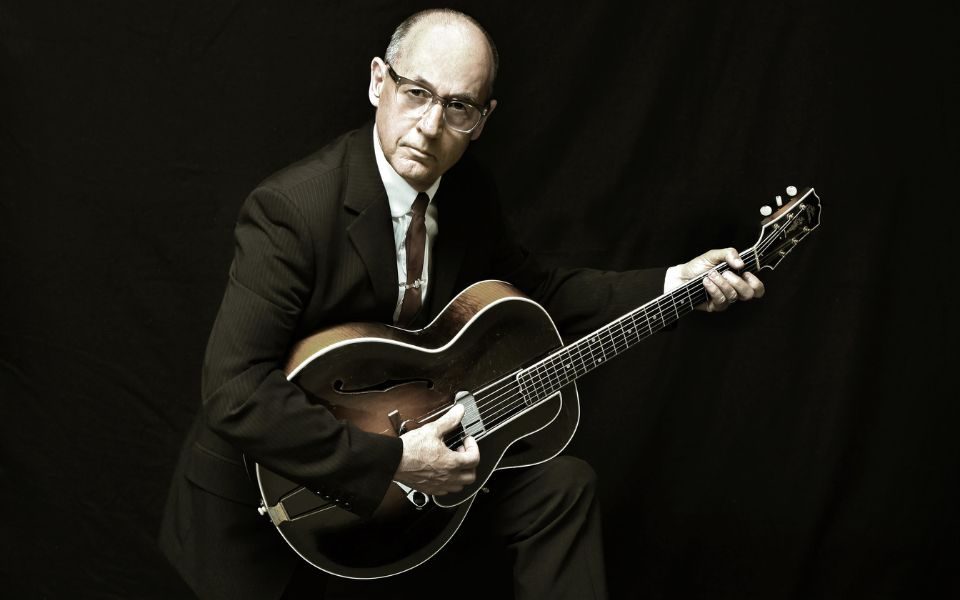 Andy Fairweather Low poses dramatically in a suit, holding an acoustic guitar against a dark, black backdrop.