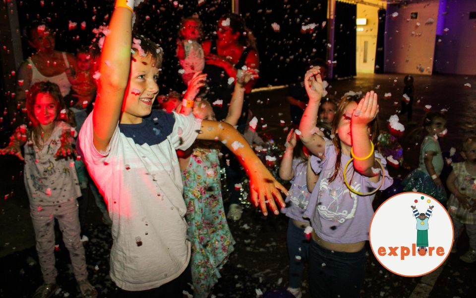 Children dance under red lights in a disco, wearing bangles and excitedly reaching out for foam falling in the air. In the bottom right corner, a roundel featuring a playful illustration of a person juggling stood on the word 'explore'.