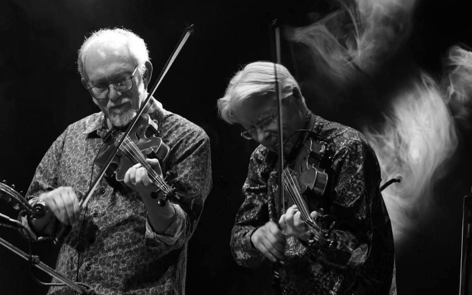 Black and white image. Bruce Molsky and Darol Anger play their violins on a low-lit, smoky stage.