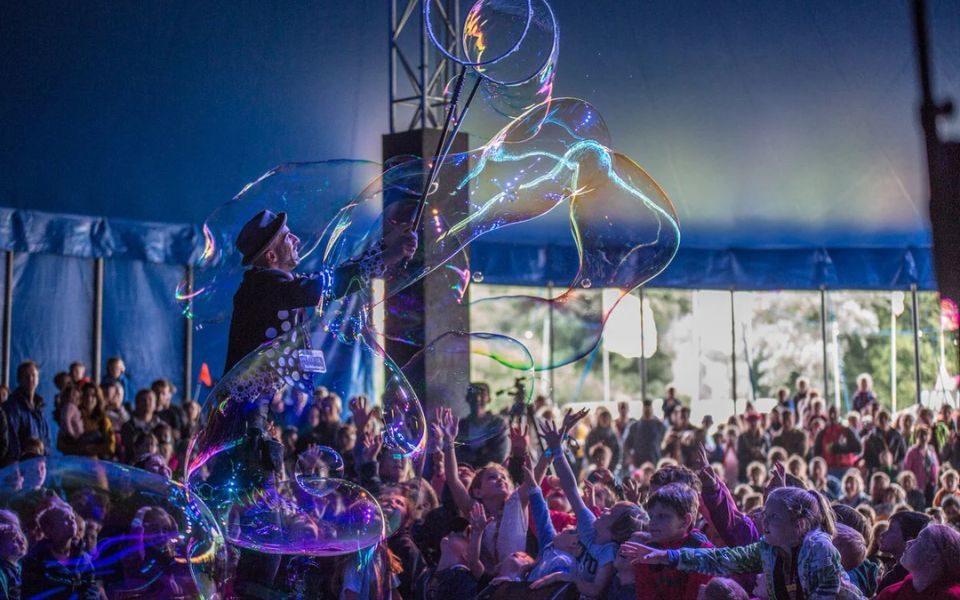 A man in a pork pie hat and polka dot waistcoat creates large bubbles on-stage in a festival tent, as children and families reach out in the audience.