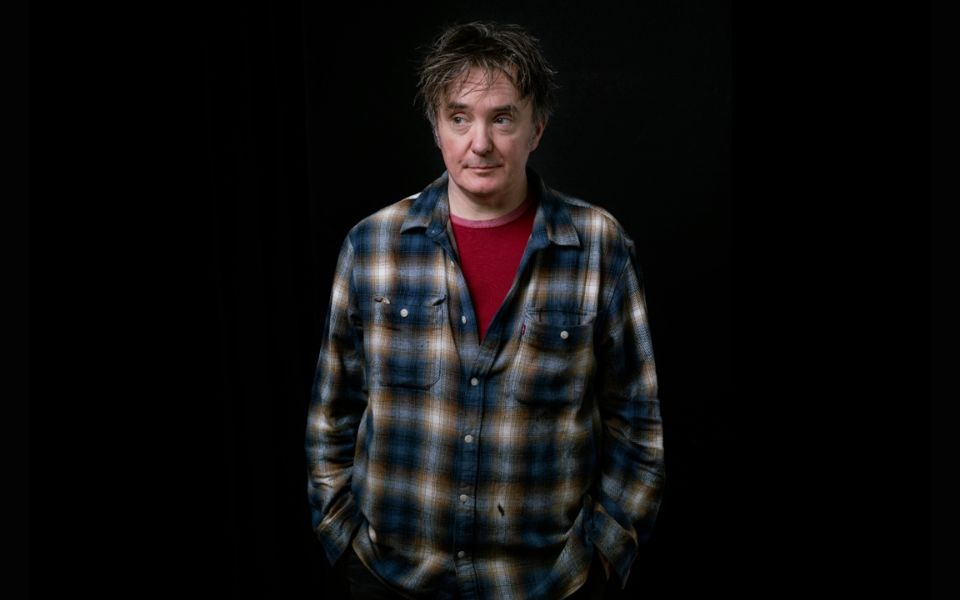 Dylan Moran stands against a black backdrop, relaxed with hands in his pockets and wearing a plaid shirt and red undershirt.