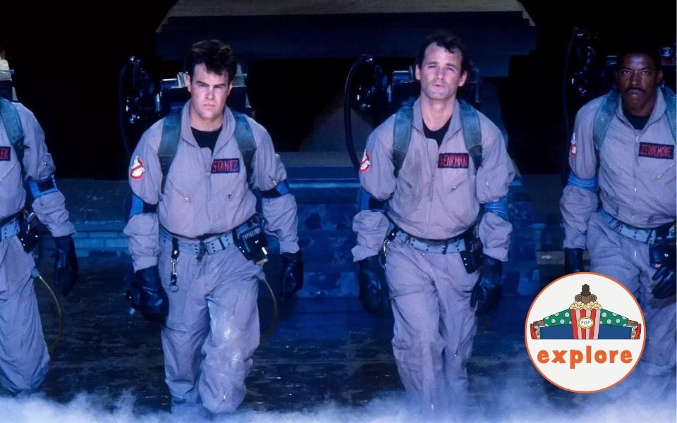 The Ghostbusters walk together from a smoky garage, in beige jumpsuits, black gloves, and technical backpacks. In the bottom right corner, a roundel featuring a playful illustration of a person sat peering over a bucket of popcorn, on the word 'explore'.