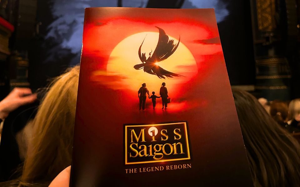 House programme for Miss Saigon with the Miss Saigon logo over the iconic artwork featuring three silhouettes walking into a red and yellow sunset beneath a black stylised phoenix.