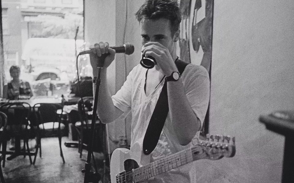 Grainy black and white still. Jeff Buckley sips from a glass covering his face, holding a microphone, with a guitar strapped round his t-shirt.