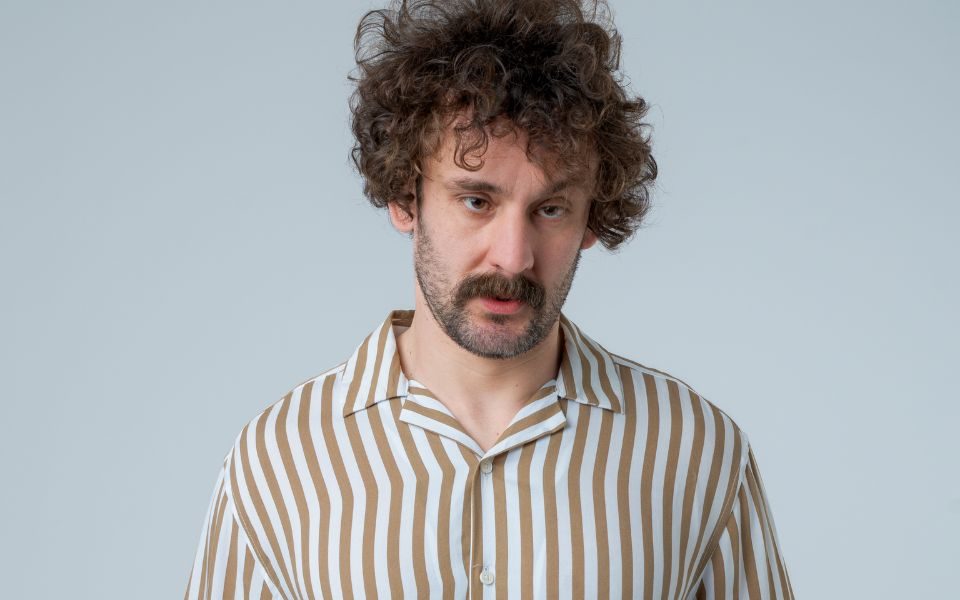 Josh Pugh stands with a neutral gaze and characteristic messy hair, wearing a striped shirt.