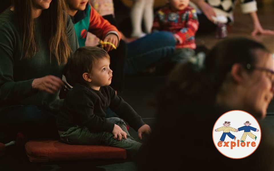 A young child sits on a cushion amongst a crowd of parents and children, looking off camera. In the bottom right corner, a roundel featuring a playful illustration of two people running across the word 'explore'.