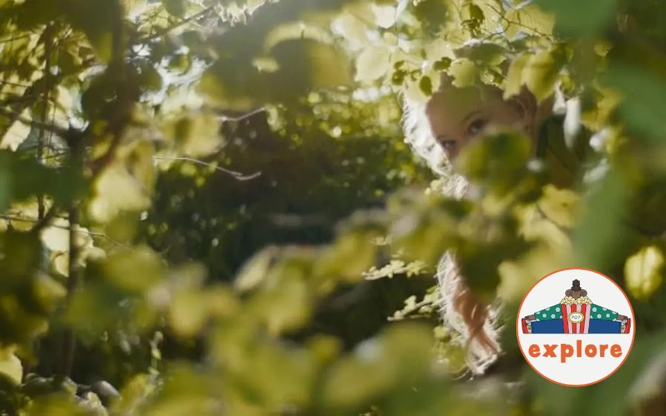 A face can be seen peeking through layers of sun-lit foliage in a green garden. In the bottom right corner, a roundel featuring a playful illustration of a person sat peering over a bucket of popcorn, on the word 'explore'.