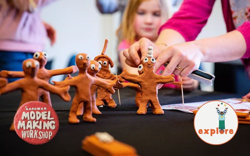 A pair of hands position an orange plasticine figurine of Morph, alongside several other Morph figurines on a black table cloth. In the bottom left corner, a clay style logo reading 'Aardman Model Making Workshop'. In the bottom right corner, a roundel featuring a playful illustration of a person juggling stood on the word 'explore'.