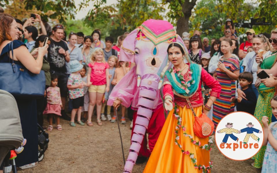 A dancer in jewellery and floral decorations performs with a person in a pink elephant costume to a crowd. In the bottom right corner, a roundel featuring a playful illustration of two people running across the word 'explore'.