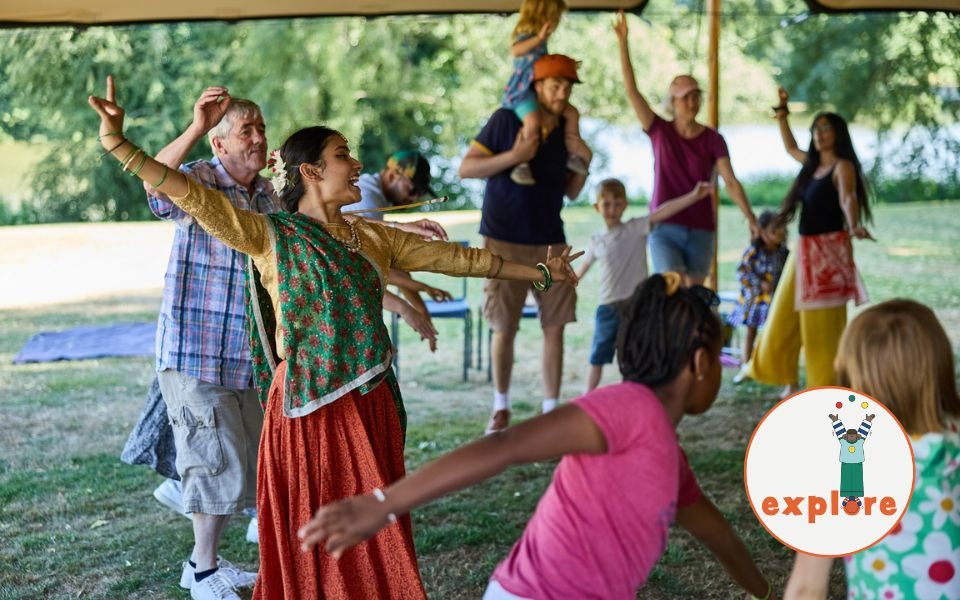 A dancer in a flowy skirt and bangles performs with a crowd of families and children, who are joining in the dance. In the bottom right corner, a roundel featuring a playful illustration of a person juggling stood on the word 'explore'.