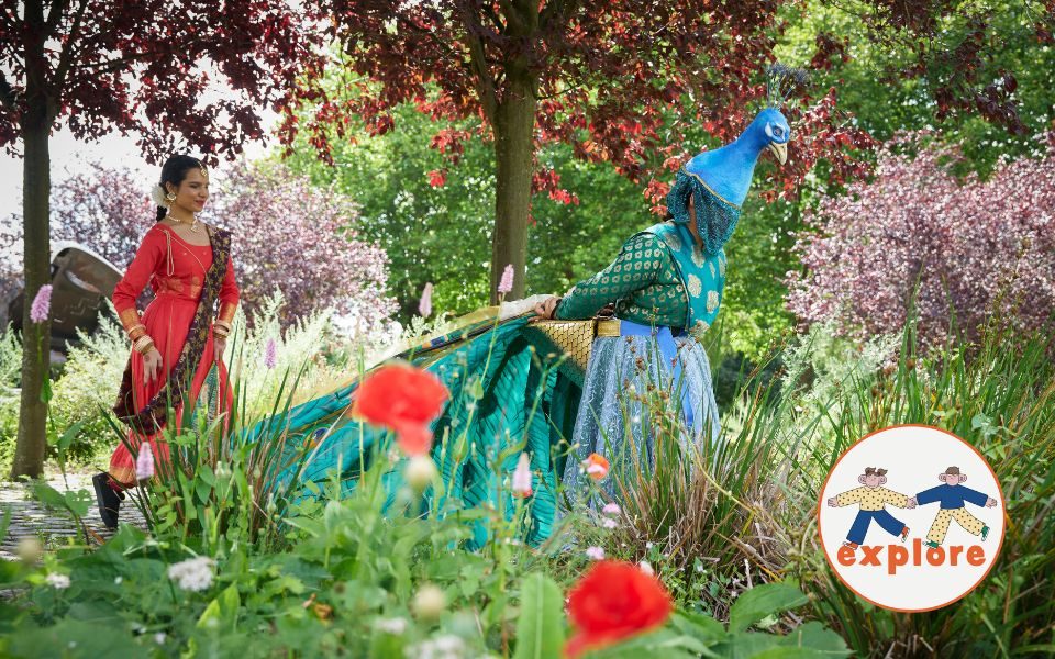 In a floral forest, a person in an elaborate peacock costume swans through the shrubbery, followed by a dancer in a red dress and gold jewellery. In the bottom right corner, a roundel featuring a playful illustration of two people running across the word 'explore'.