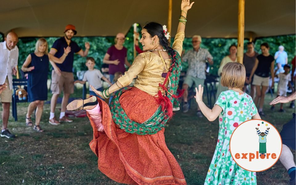 A dancer in a flowy skirt and bangles performs to a crowd, alongside a child, who are mimicking the dance. In the bottom right corner, a roundel featuring a playful illustration of a person juggling stood on the word 'explore'.