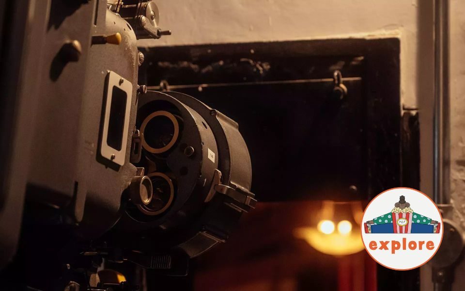 A projector in a cinema projection booth. In the bottom right corner, a roundel featuring a playful illustration of a person sat peering over a bucket of popcorn, on the word 'explore'.