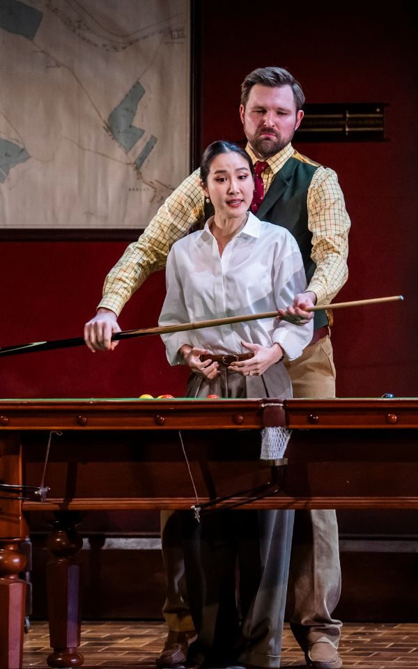 James Newby as Count Almaviva standing with his arms around Hera Hyesang Park to hold a snooker cue in front of her by a snooker table.