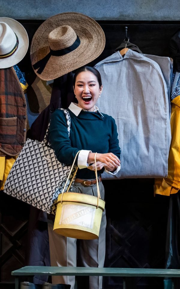 Hera Hyesang Park laughing as Susanna carrying a hat box and large handbag in fron of lots of clothes and hats hung on hooks.