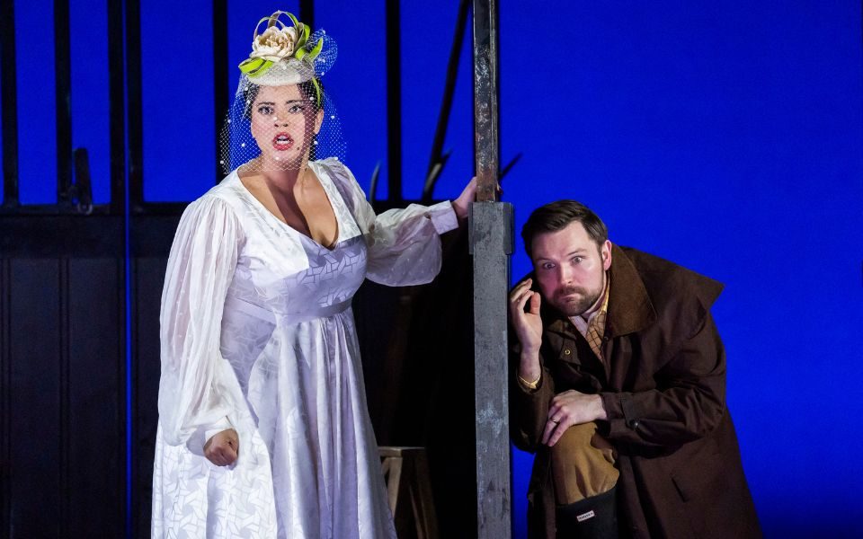 A woman in a white wedding dress and flower fascinator looks out at the audience. A man crouches to hide on the other side of the fence she holds.