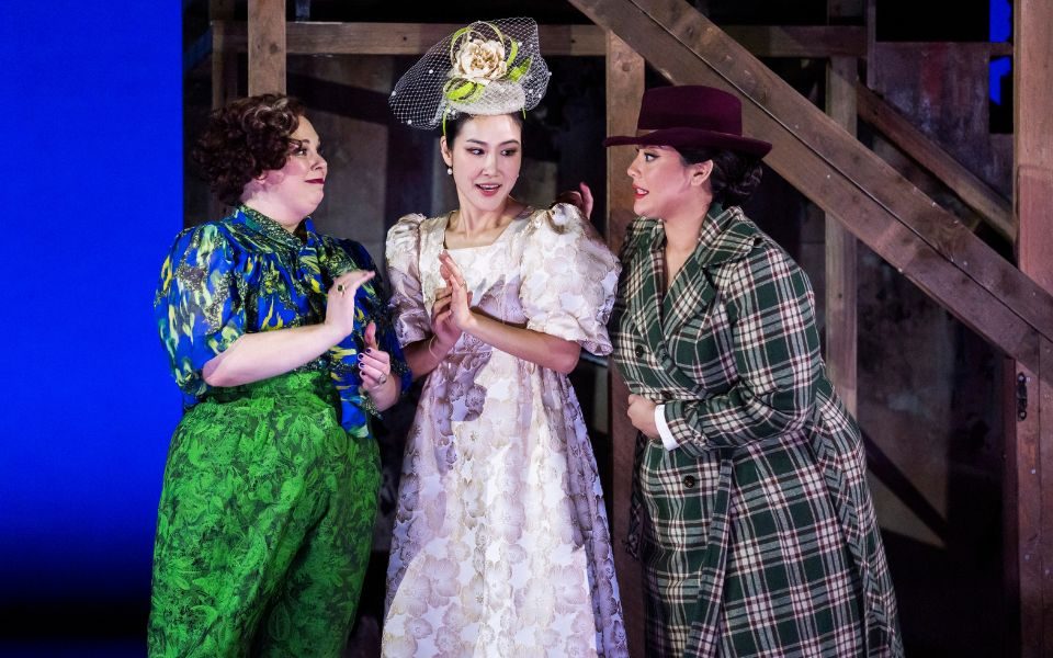 Three women talking to each in formal dress. The one on the left wears a blue and green dress, the one in the middle wears a pale pink dress and fascinator on her head, and the one on the right wears a long green check coat and brown hat.
