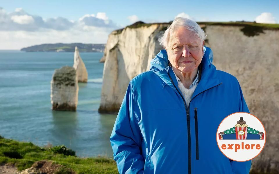 David Attenborough stands at a cliff edge with sea visible in the background, facing the camera in a blue coat. In the bottom right corner, a roundel featuring a playful illustration of a person sat peering over a bucket of popcorn, on the word 'explore'.