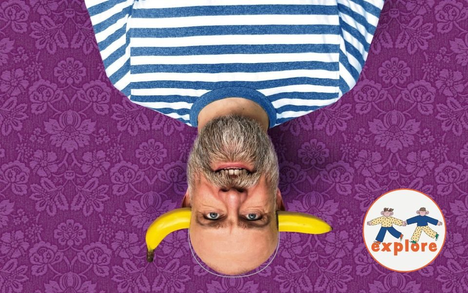 Upside down, Olaf Falafel grins at the camera, wearing a prop banana headband and stripy blue and white t-shirt, positioned over a patterned purple background. In the bottom right corner, a roundel featuring a playful illustration of two people running across the word 'explore'.