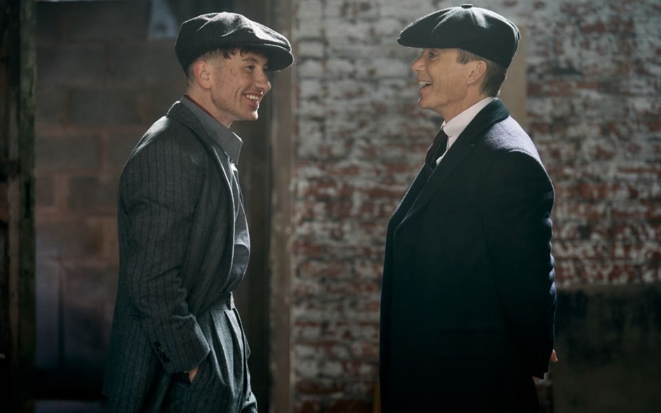 Cillian Murphy and Barry Keoghan smile, both in profile wearing period style suits and long coats with flat caps.