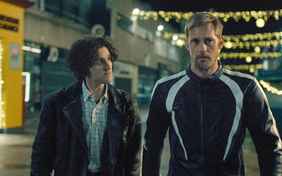 Alexander Skarsgård in a biker jacket and Harry Melling in a leather jacket walk together through a street at night.