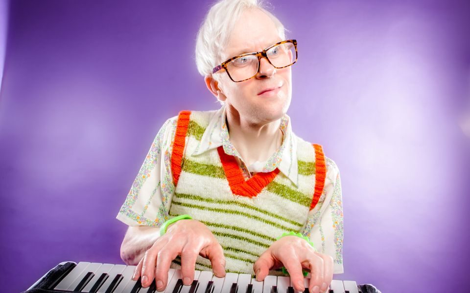 Robert White in glasses and a sweater vest over a patterned shirt, sat at a keyboard in front of a purple backdrop.