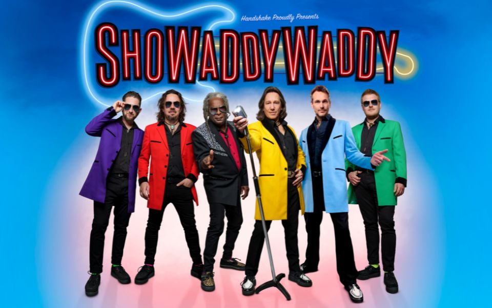 The members of Showaddywaddy stand in brightly coloured suit jackets, posing in sunglasses against a vibrant blur backdrop. In front of a neon guitar sign, text reads: Handshake Proudly Presents Showaddywaddy.